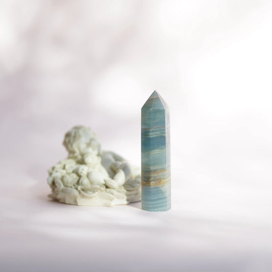 A polished blue onyx crystal point tower with horizontal banding stands upright next to a small white sculptural figurine on a soft white surface against a pale background.