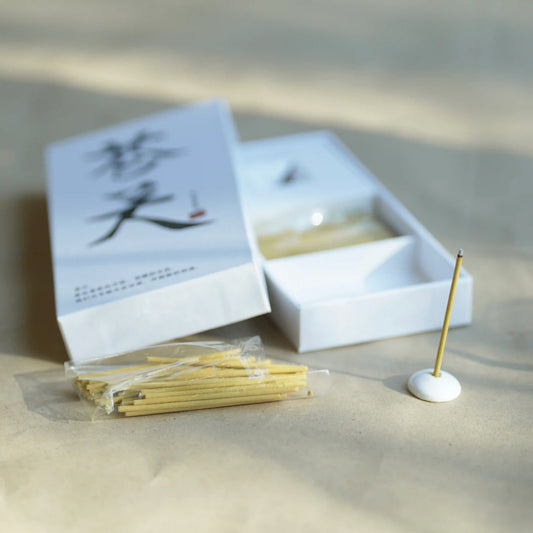 A set of incense sticks in a white box with a white incense holder, photographed on a beige surface with soft natural shadows.