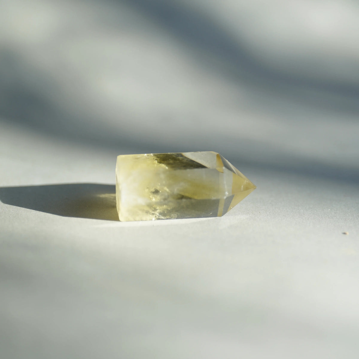 A natural yellow quartz crystal point with internal inclusions, resting on a light gray surface under direct sunlight with soft shadows.
