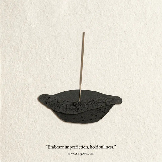A minimalist black mochi mochi incense holder with a single vertical incense stick inserted, set against a textured off-white background. The text 'Embrace imperfection, hold stillness.