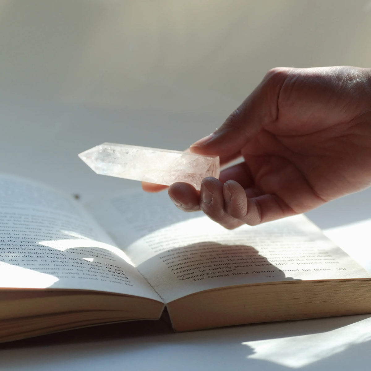 A hand holds a clear quartz crystal point over an open book with soft sunlight filtering in from the side.