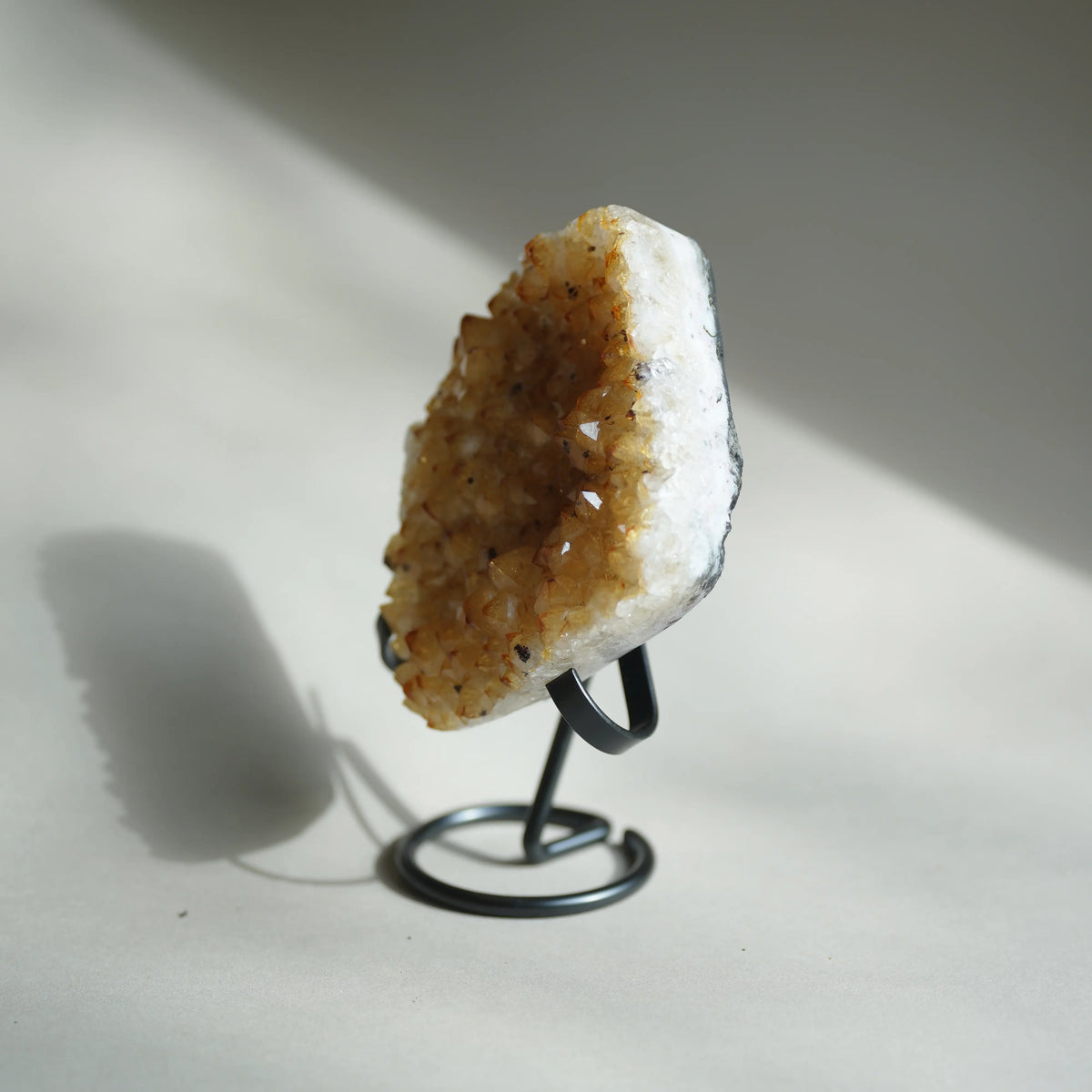 A natural crystal geode cluster with golden-brown druzy crystals and a white matrix, displayed on a black metal stand against a neutral beige background with soft shadows.