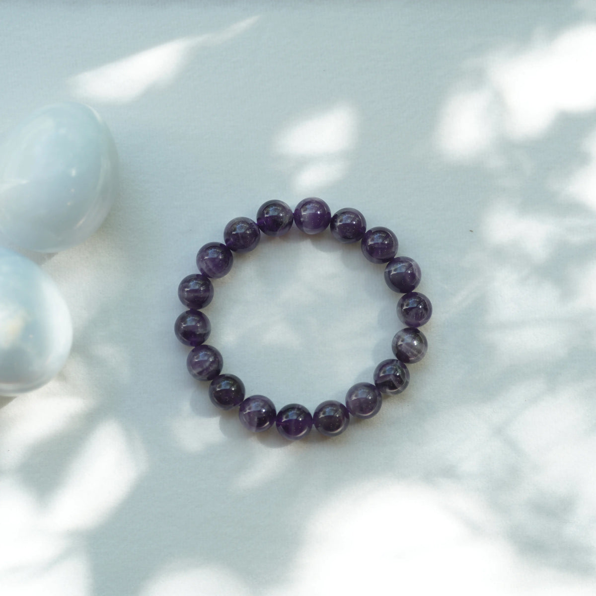 A beaded stretch bracelet made of polished dark purple amethyst round beads, displayed on a light gray surface with soft leaf shadows and two white stones in the background.