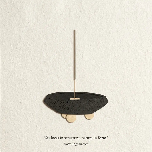 A minimalist black speckled stone bowl pendant with a thin brass rod and small wooden feet, mounted on a textured off-white wall. Text below reads 'Stillness in structure, nature in form.