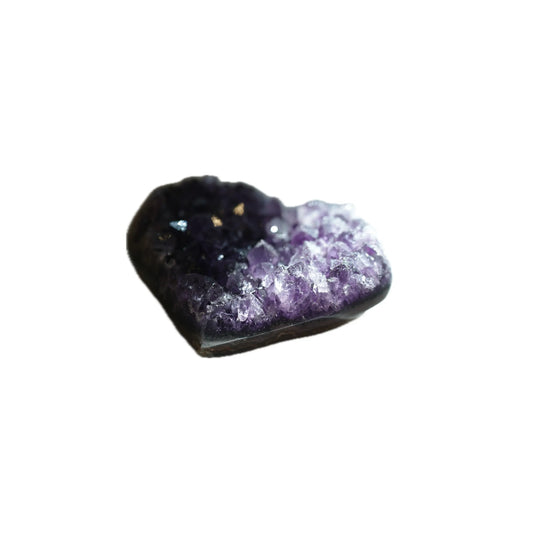 A slightly angled profile view of the same heart-shaped Amethyst crystal geode, emphasizing its depth and organic form. Natural light dramatically illuminates the sparkling purple and clear crystalline formations in the right foreground, while the background remains grounded in shadow, curated for manifestation and grounding by Singoau.