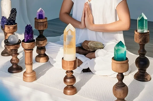 A person in a white outfit sits cross-legged with hands in prayer pose, surrounded by wooden crystal holders displaying various colored gemstones and crystals on a white surface.