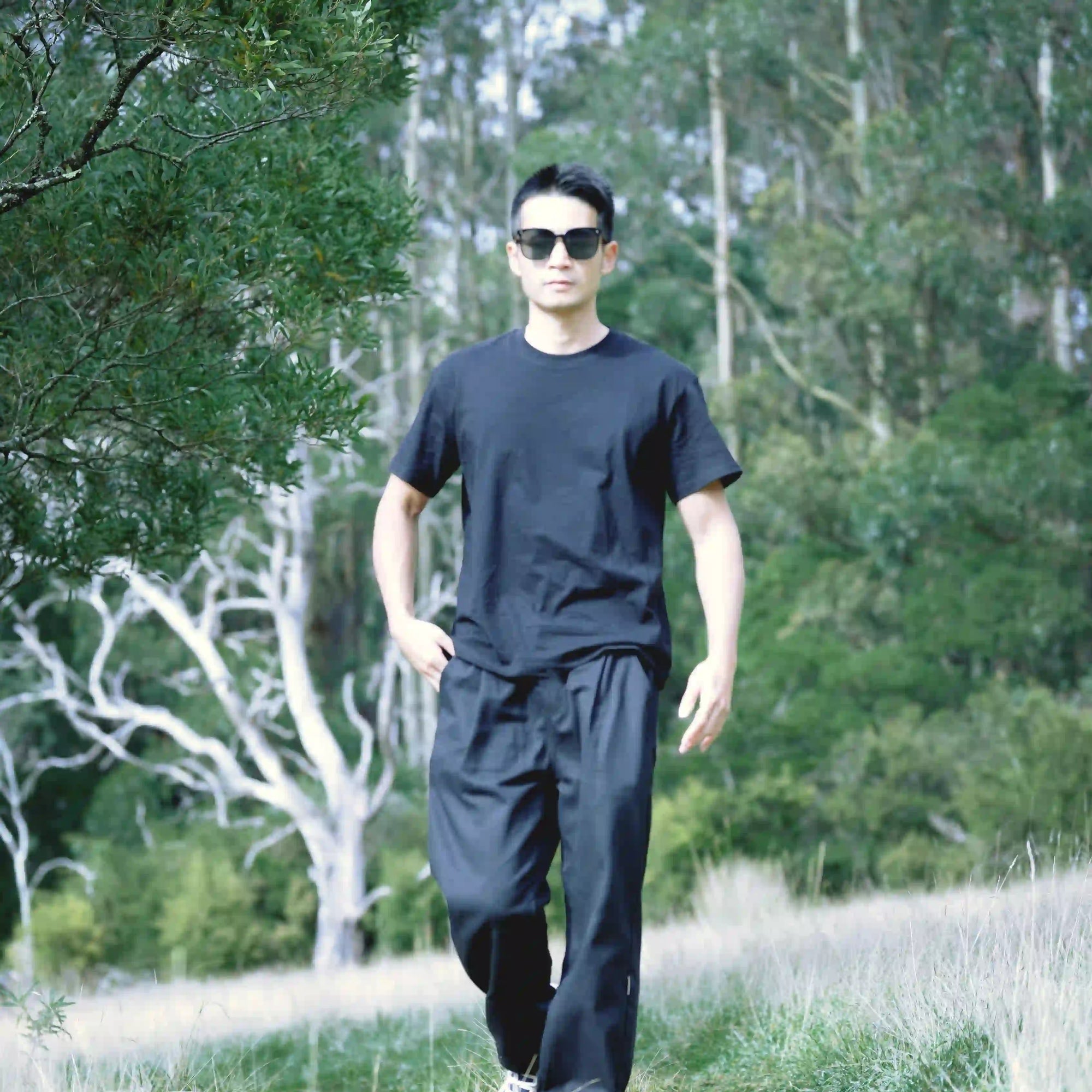 A man wearing a black t-shirt and black sweatpants walking outdoors in a grassy field with trees in the background.