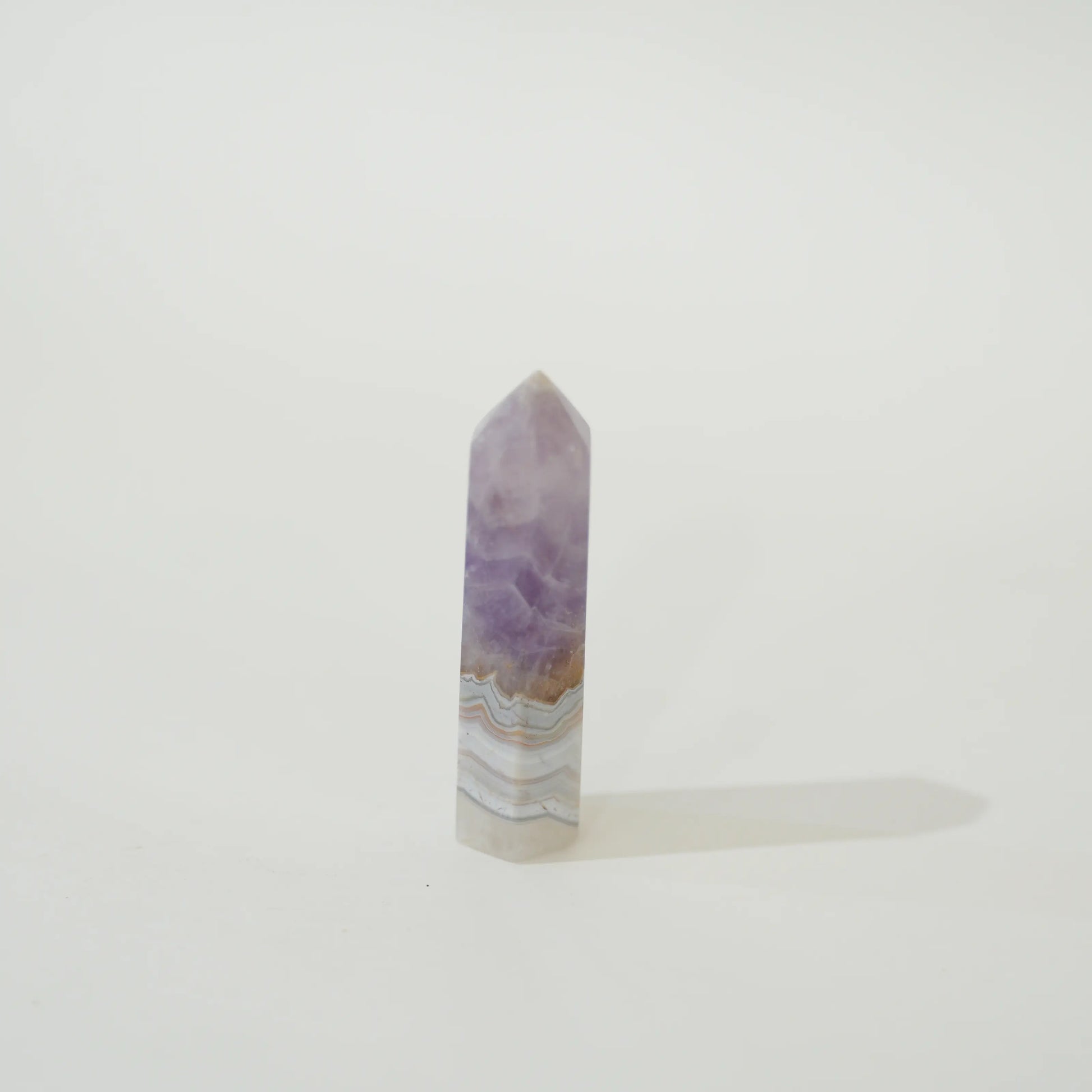 A polished Amethyst & Mexican Lace Agate Tower with natural banding of purple, white, and light brown, standing upright on a white surface against a white background.