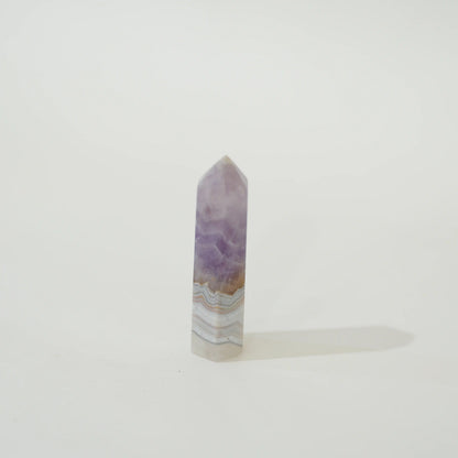 A polished Amethyst & Mexican Lace Agate Tower with natural banding of purple, white, and light brown, standing upright on a white surface against a white background.