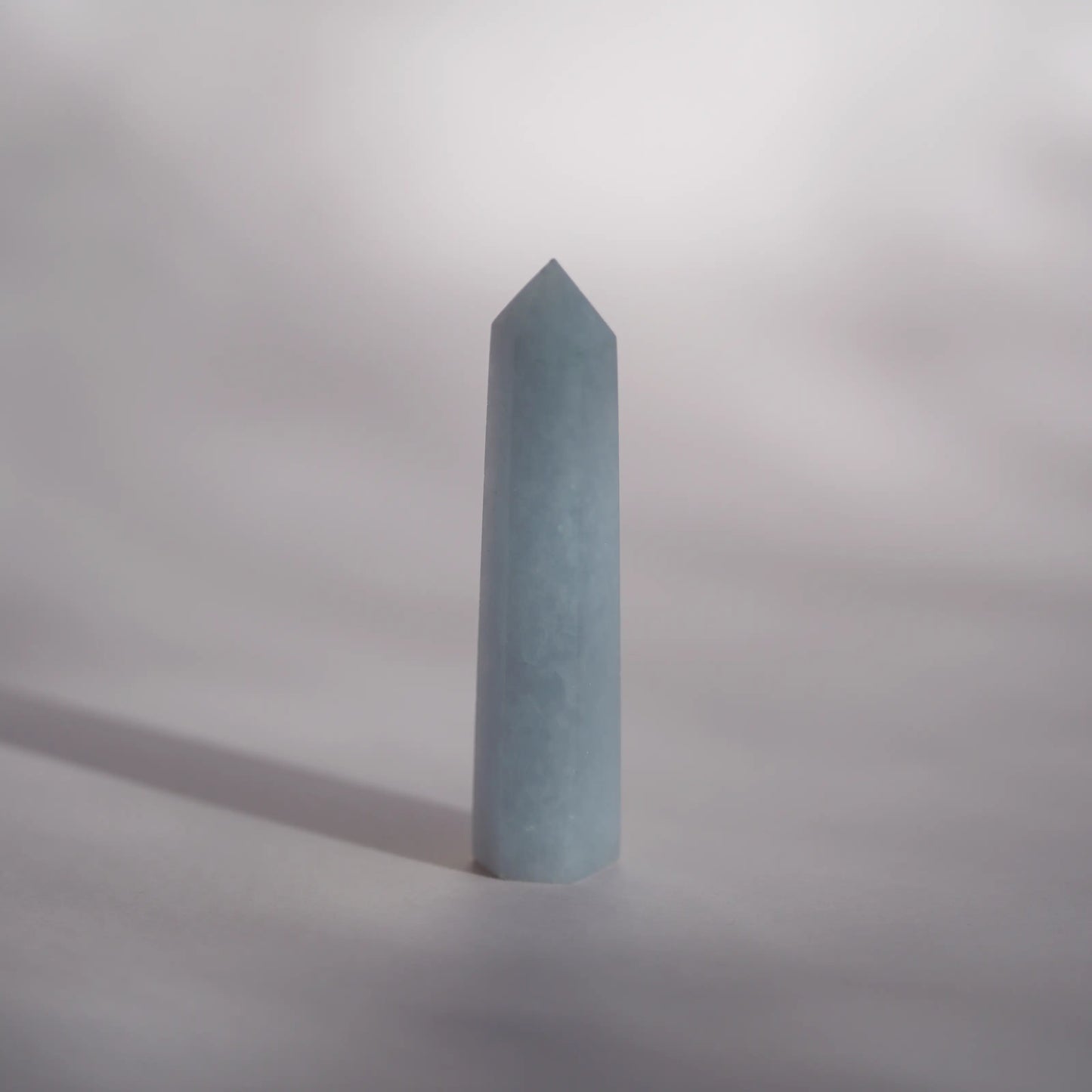 A celestite crystal tower or obelisk standing upright on a neutral gray surface with a soft shadow cast to the left.