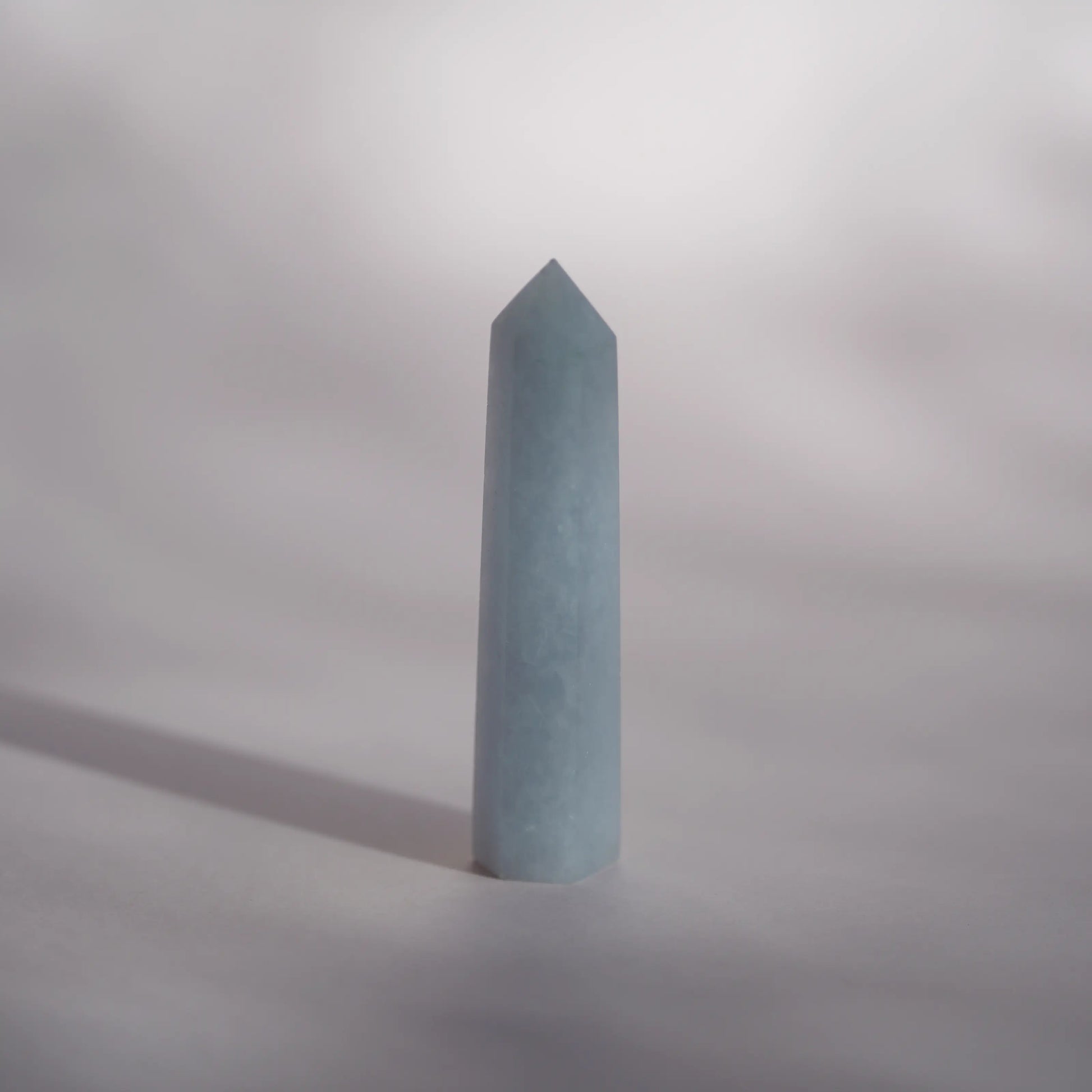 A celestite crystal tower or obelisk standing upright on a neutral gray surface with a soft shadow cast to the left.
