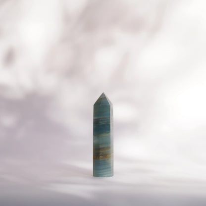 A polished blue onyx crystal point tower with horizontal banding, standing upright on a soft white surface against a blurred light background.