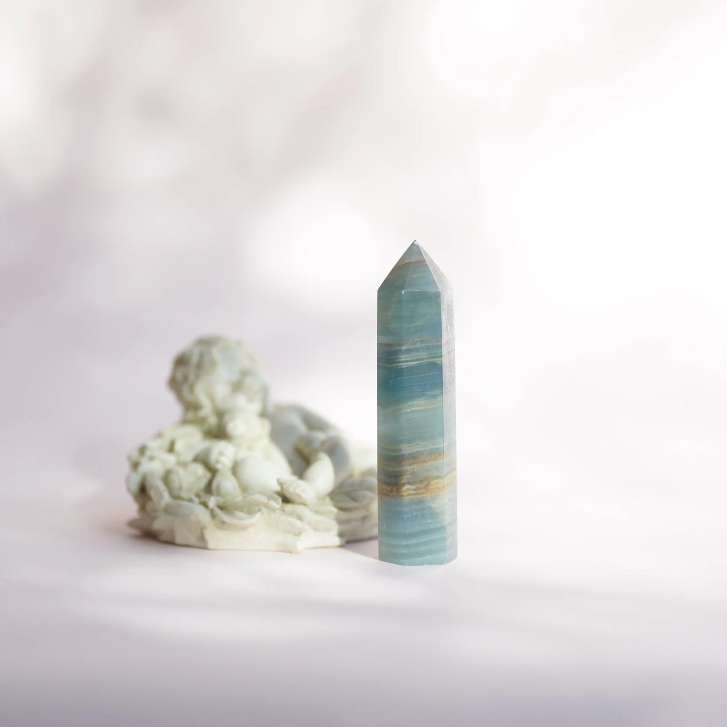 A polished blue onyx crystal point tower  with horizontal banding stands upright next to a small white sculptural figurine on a soft white surface against a pale background.