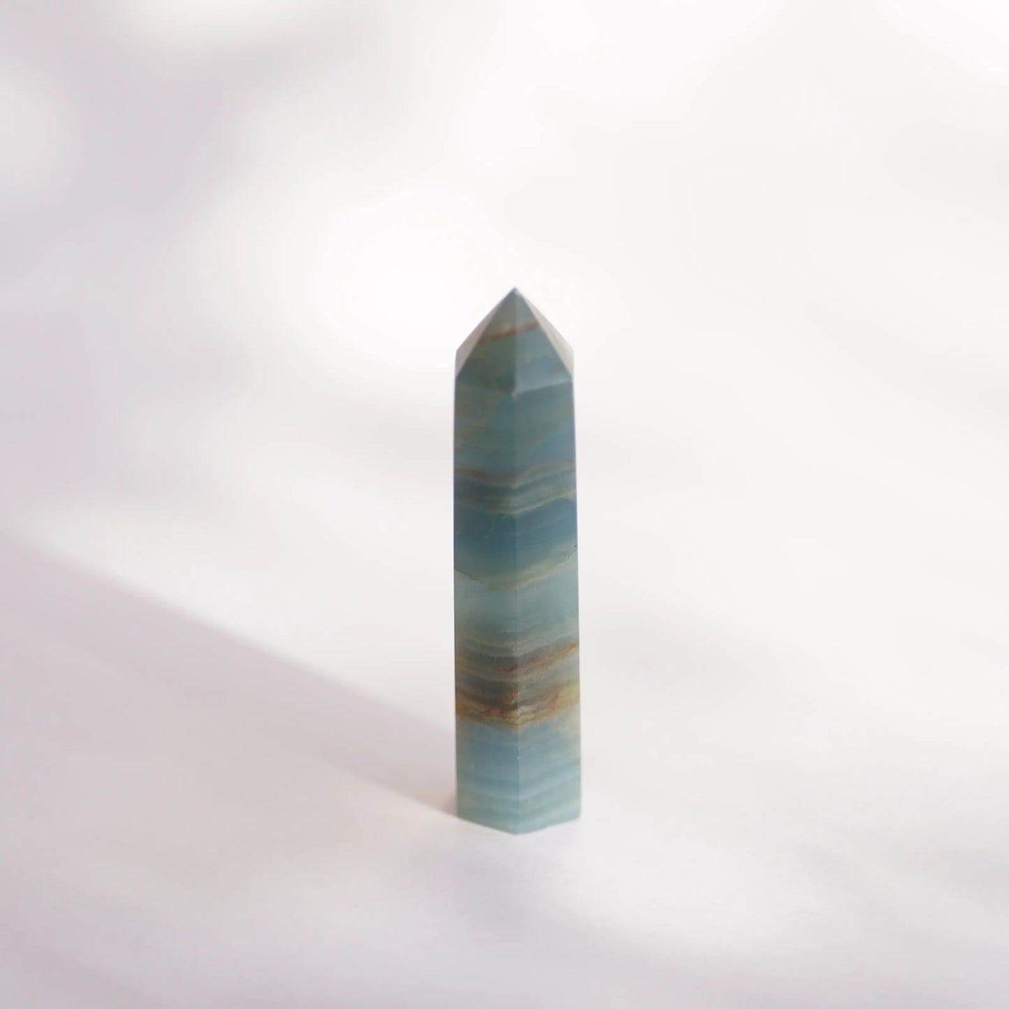 A polished blue onyx crystal point tower , likely amazonite, standing upright on a white surface with a soft shadow cast to the left.