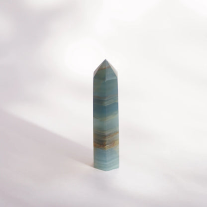 A polished blue onyx crystal point tower , likely amazonite, standing upright on a white surface with a soft shadow cast to the left.