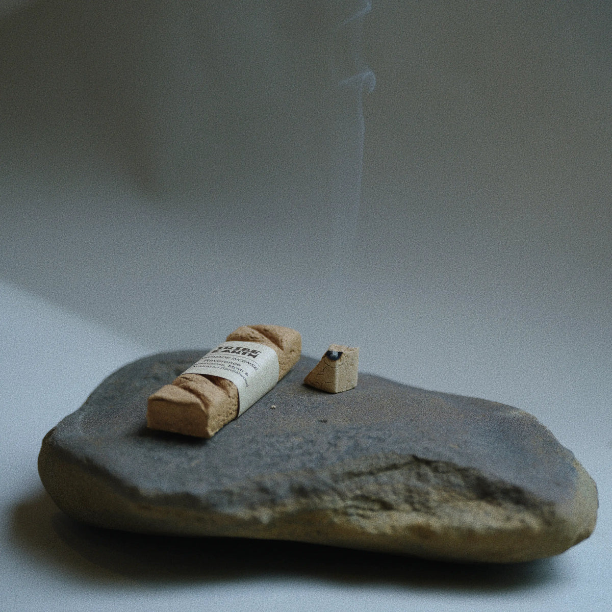 Tribe earth incense stands beside a labeled cork-like burning incense with smoke log on a dark stone slab.