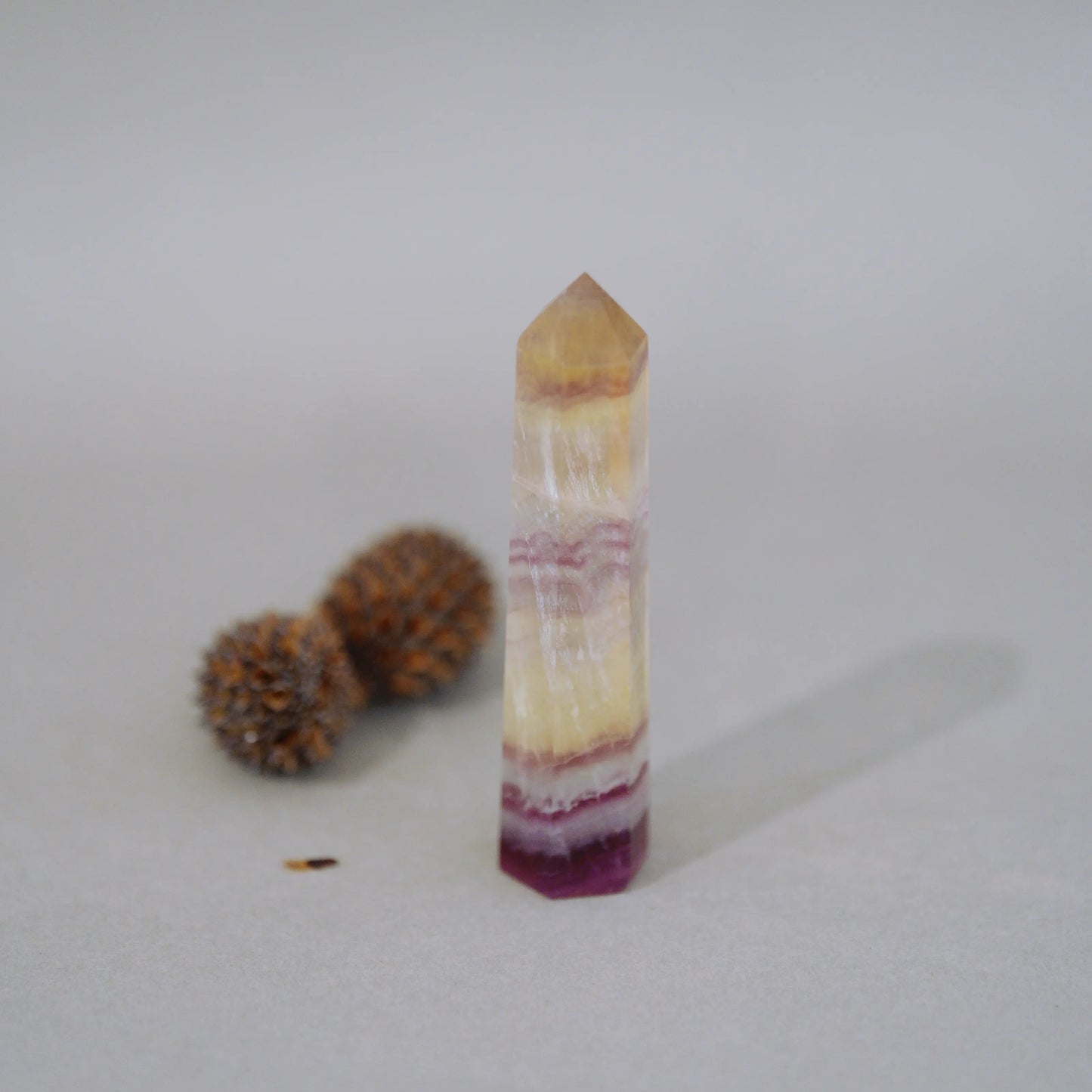 A natural Candy Fluorite Tower with horizontal banding in shades of yellow, cream, pink, and deep purple, standing upright on a light gray surface with two small brown decorative balls in the background.