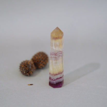 A natural Candy Fluorite Tower with horizontal banding in shades of yellow, cream, pink, and deep purple, standing upright on a light gray surface with two small brown decorative balls in the background.