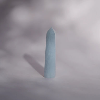 A pale blue celestite tower standing upright on a soft gray surface with a soft shadow cast to the left.