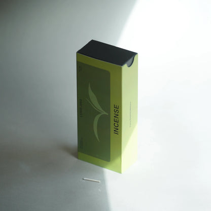 A rectangular incense box with a lime green and dark green front panel, black top, and vertical 'INCENSE' text, standing on a light gray surface with a single incense stick in front.