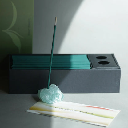 A teal incense stick stands upright in a light aqua crystal holder, with a dark charcoal incense box behind it and a printed card in front on a gray surface.