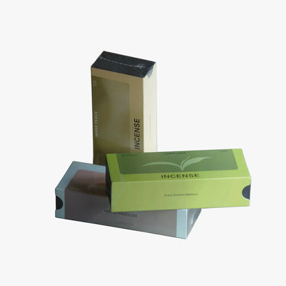Three product boxes for incense are arranged on a neutral gray studio background. A tall beige box stands upright in the back, a bright green box labeled 'INCENSE' rests in front, and a gray box with blue edges lies underneath.