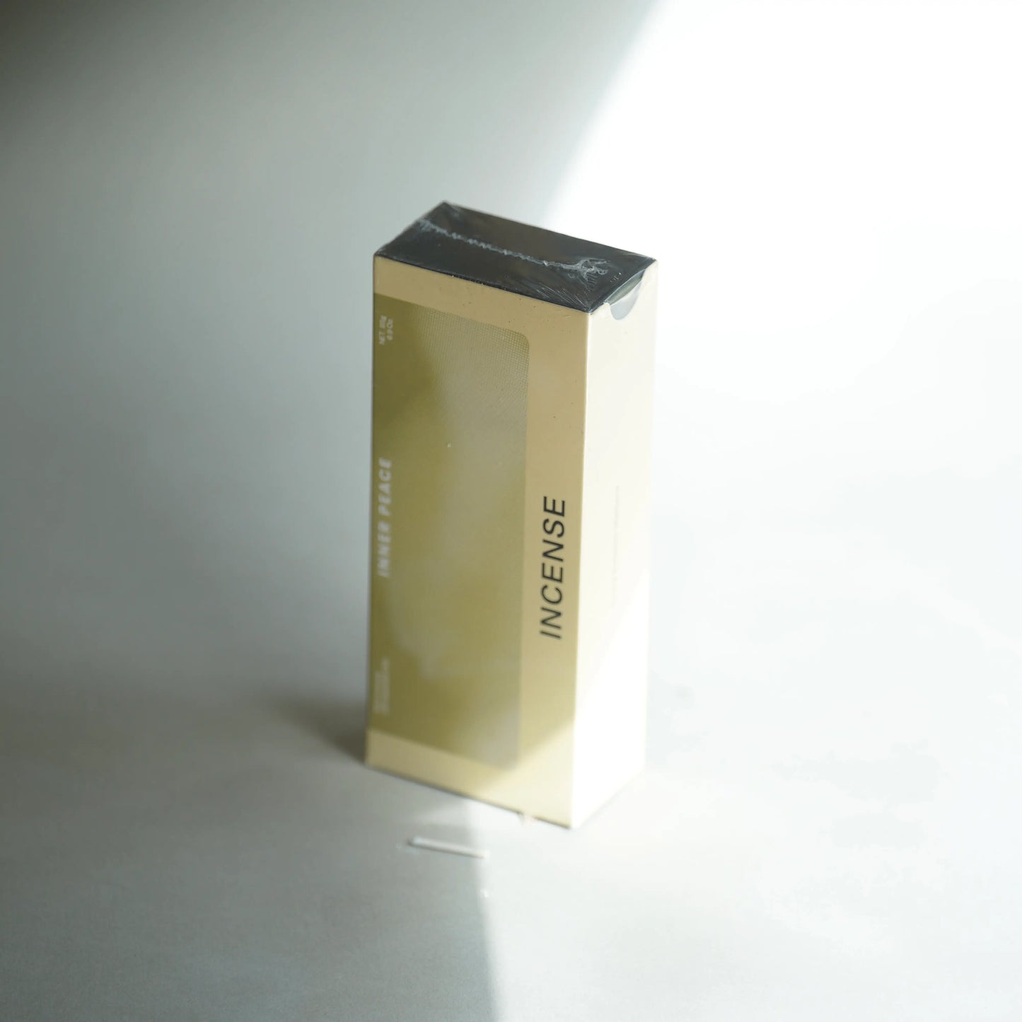 A rectangular product box for incense, standing upright on a light gray surface. The box is a warm beige/cream color with a transparent window on the front, black text reading 'INNER PEACE' and 'INCENSE', and a dark gray top surface.