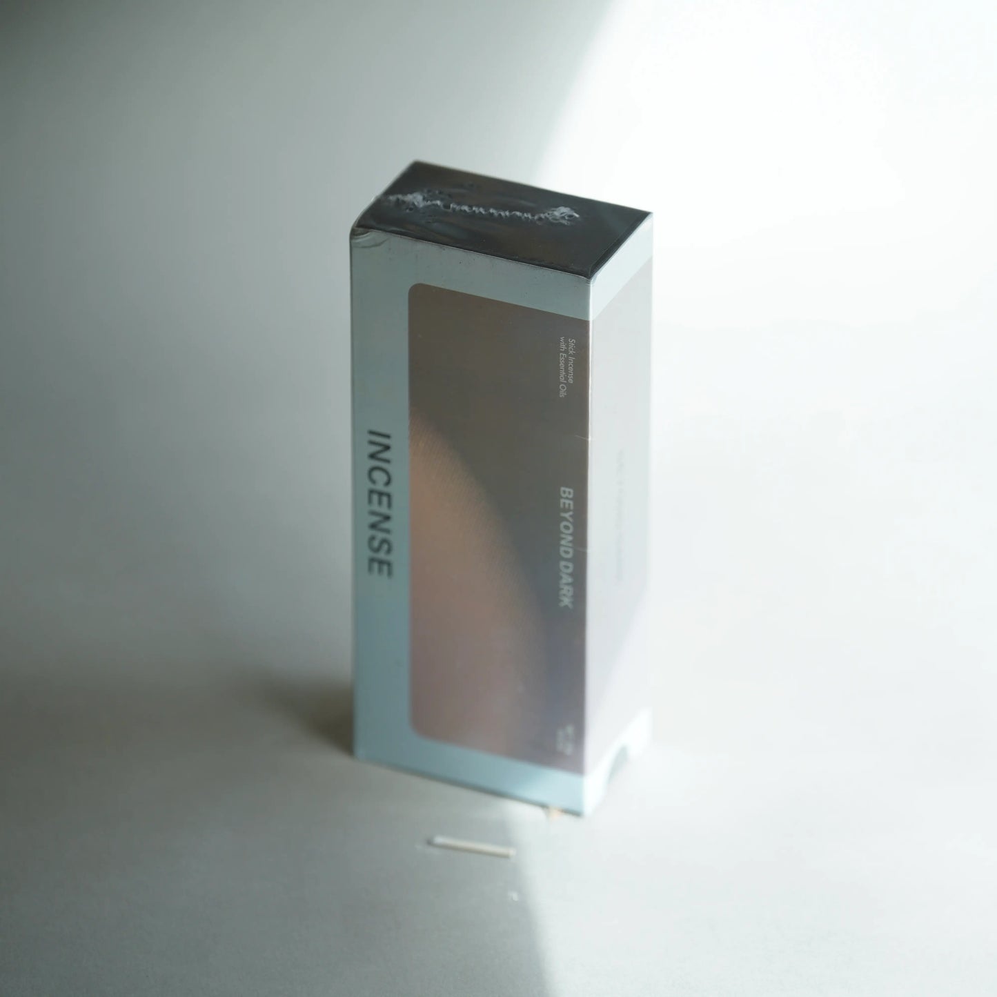 A rectangular incense box standing upright on a light gray surface. The box is mostly white and light gray with a large dark brownish-gray front panel and black top flap.