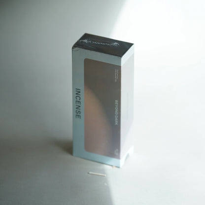 A rectangular incense box standing upright on a light gray surface. The box is mostly white and light gray with a large dark brownish-gray front panel and black top flap.
