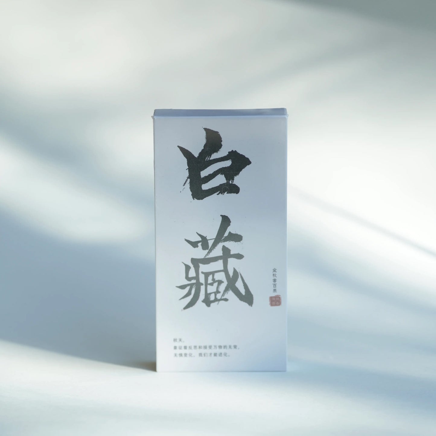 A tall, narrow white product box with large black Chinese calligraphy and small red seal details, standing upright on a softly lit white surface with natural shadows.