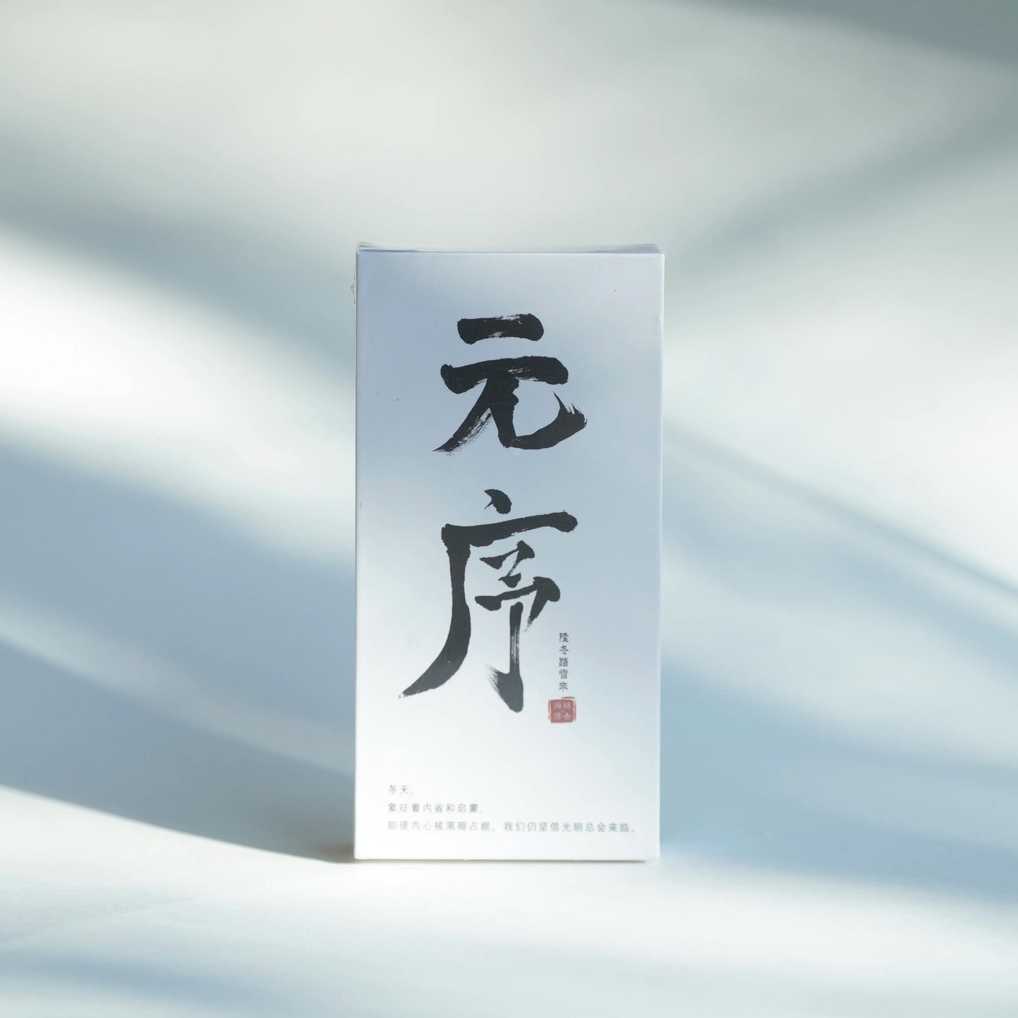 A tall, narrow white product box with large black Chinese calligraphy on the front and a small red seal, standing upright on a softly lit white surface with dappled shadows.