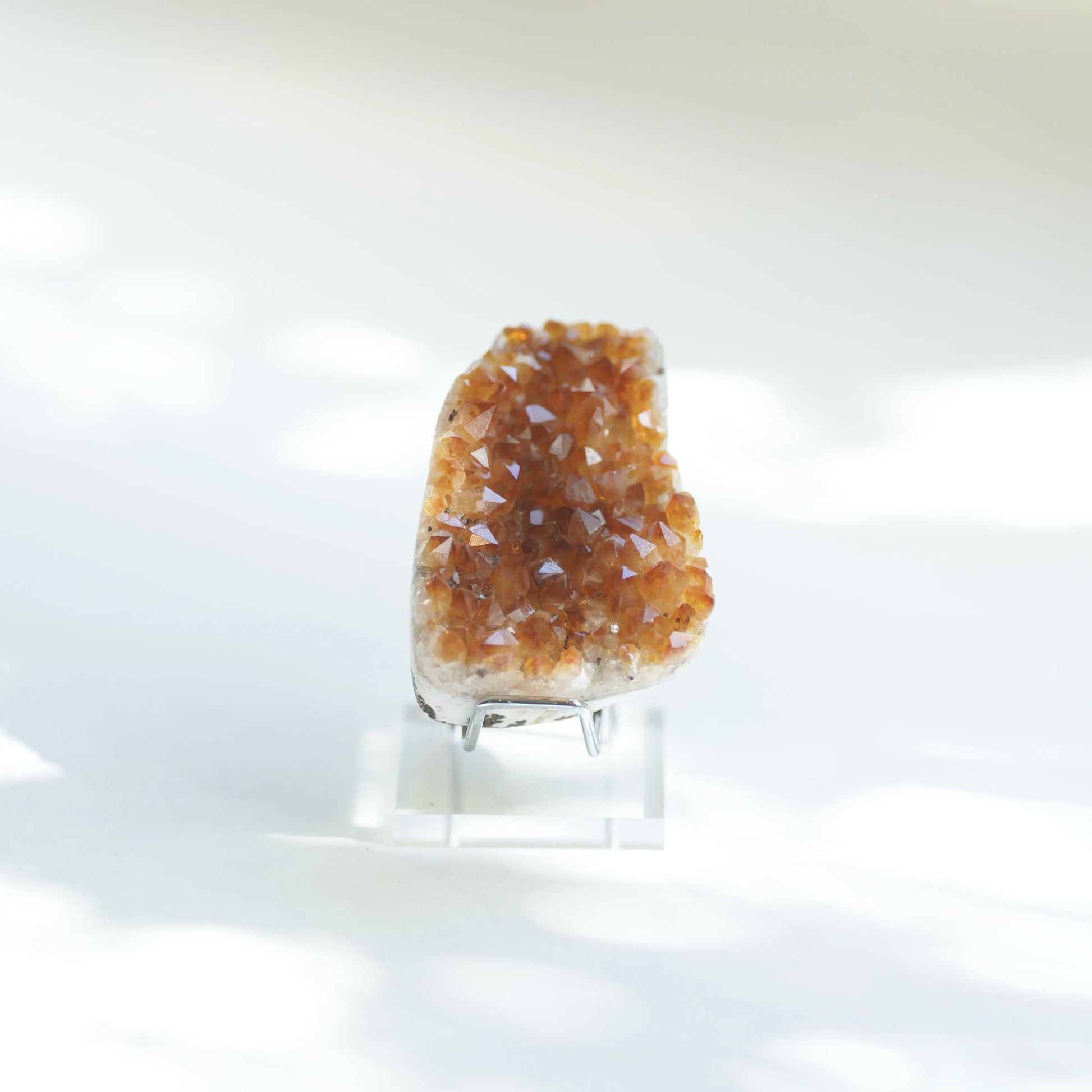 A natural crystal geode specimen with orange-brown druzy crystals on a clear acrylic display stand, photographed against a bright white background with soft light shadows.