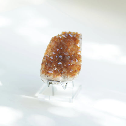 A citrine crystal cluster with brown and amber-colored crystalline points, mounted on a clear acrylic stand against a bright white background.