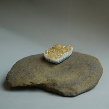A small polished crystal or gemstone with a white base and golden-yellow glittery top, displayed on a large natural stone against a neutral gray background.