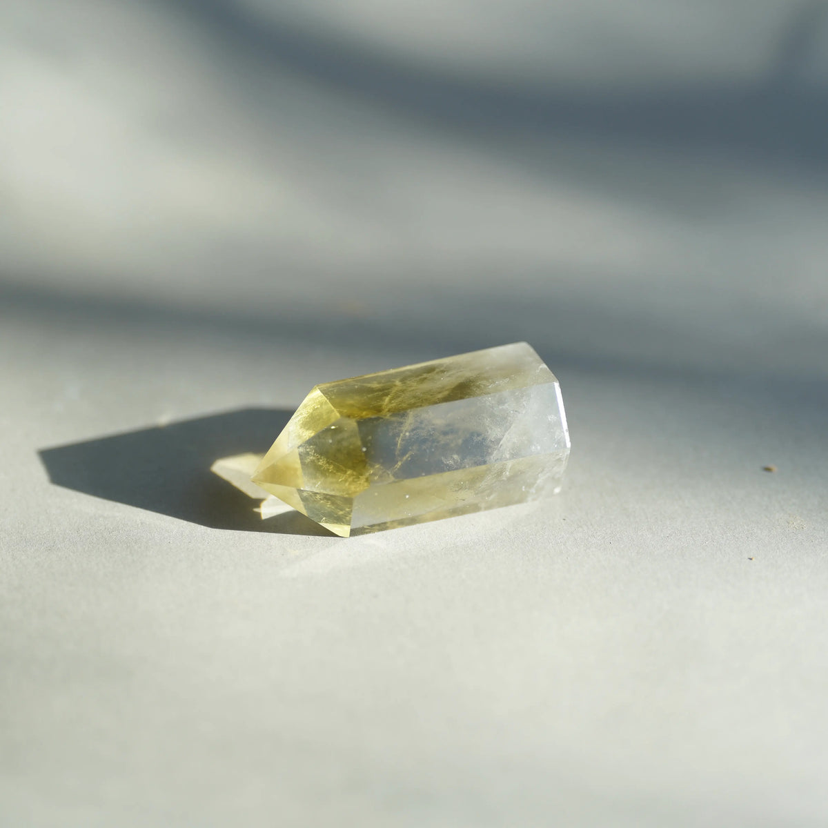 A faceted clear crystal with a pale yellow tint, resting on a neutral gray surface under direct sunlight that casts a sharp shadow.