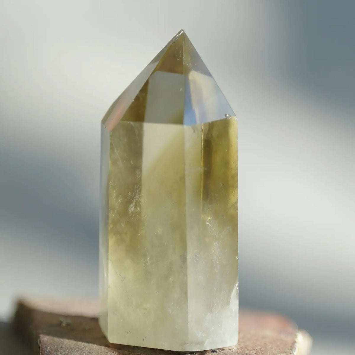 A natural pointed crystal tower, likely quartz, showing a translucent yellow-green body with a clear faceted top, resting on a small stone surface against a soft, blurred gray background.