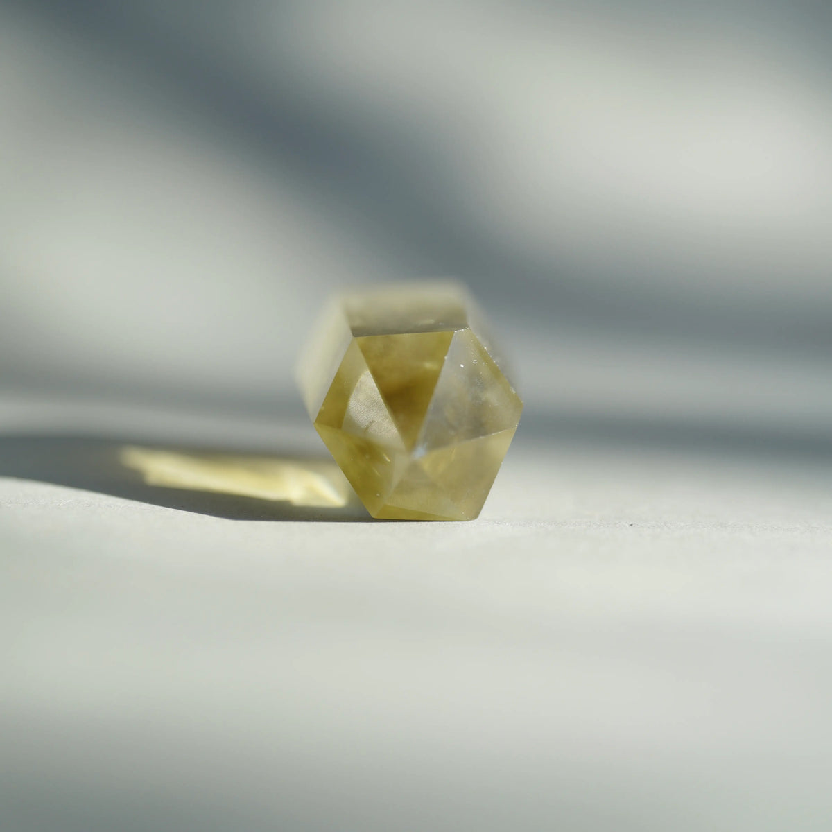 A faceted crystal sphere, likely a citrine or yellow quartz gemstone, resting on a light gray surface with soft shadows and a blurred background.