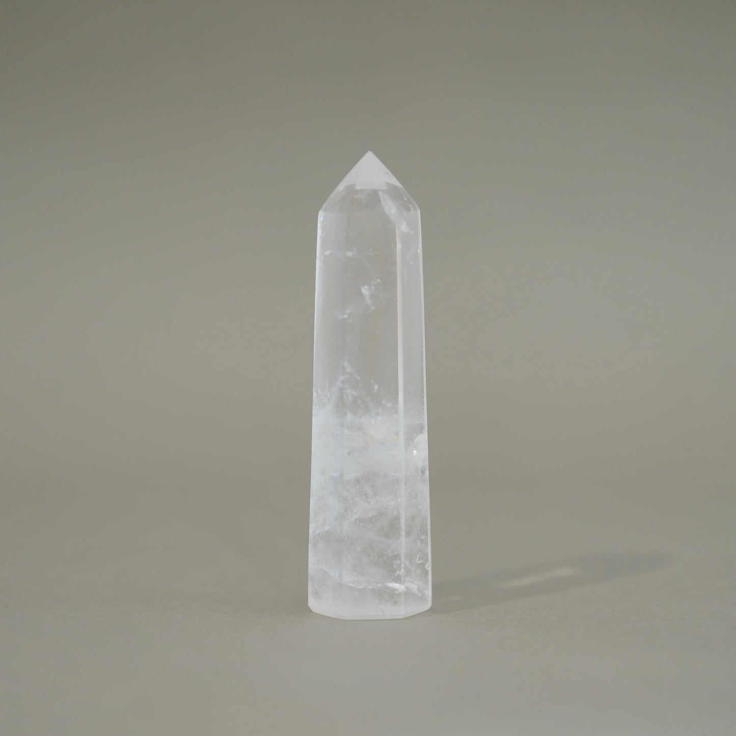 A natural clear quartz crystal point tower with internal inclusions, standing upright on a neutral gray background.