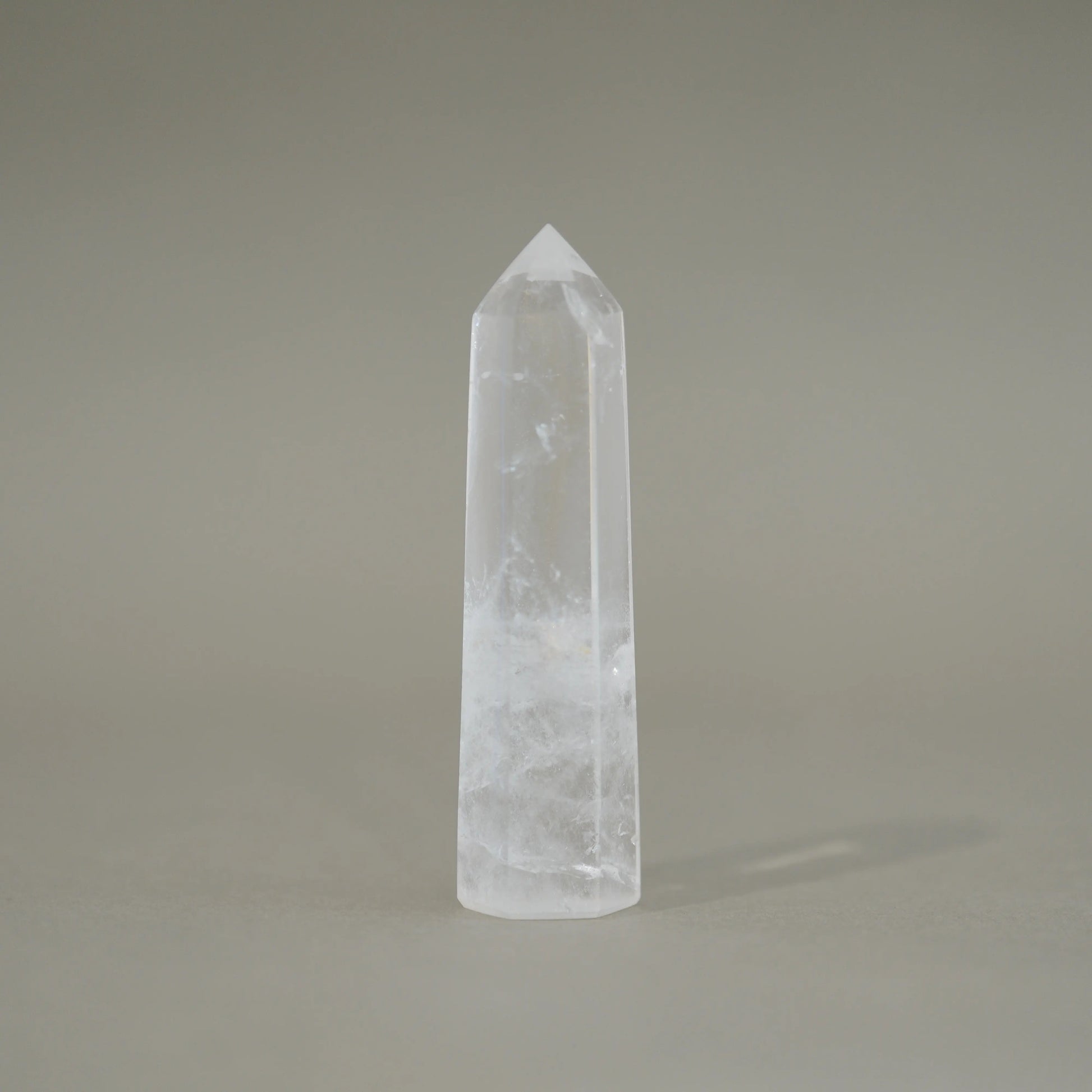 A natural clear quartz crystal point tower with internal inclusions, standing upright on a neutral gray background.