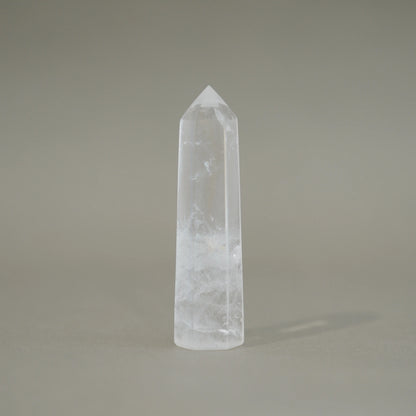 A natural clear quartz crystal point tower with internal inclusions, standing upright on a neutral gray background.