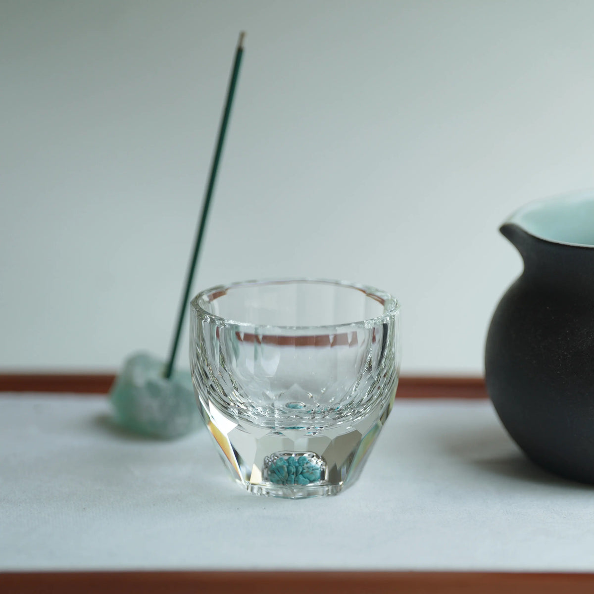 Close-up of a crystal tumbler with a black straw, turquoise garnish, and a black tea mug on a white tea plate.