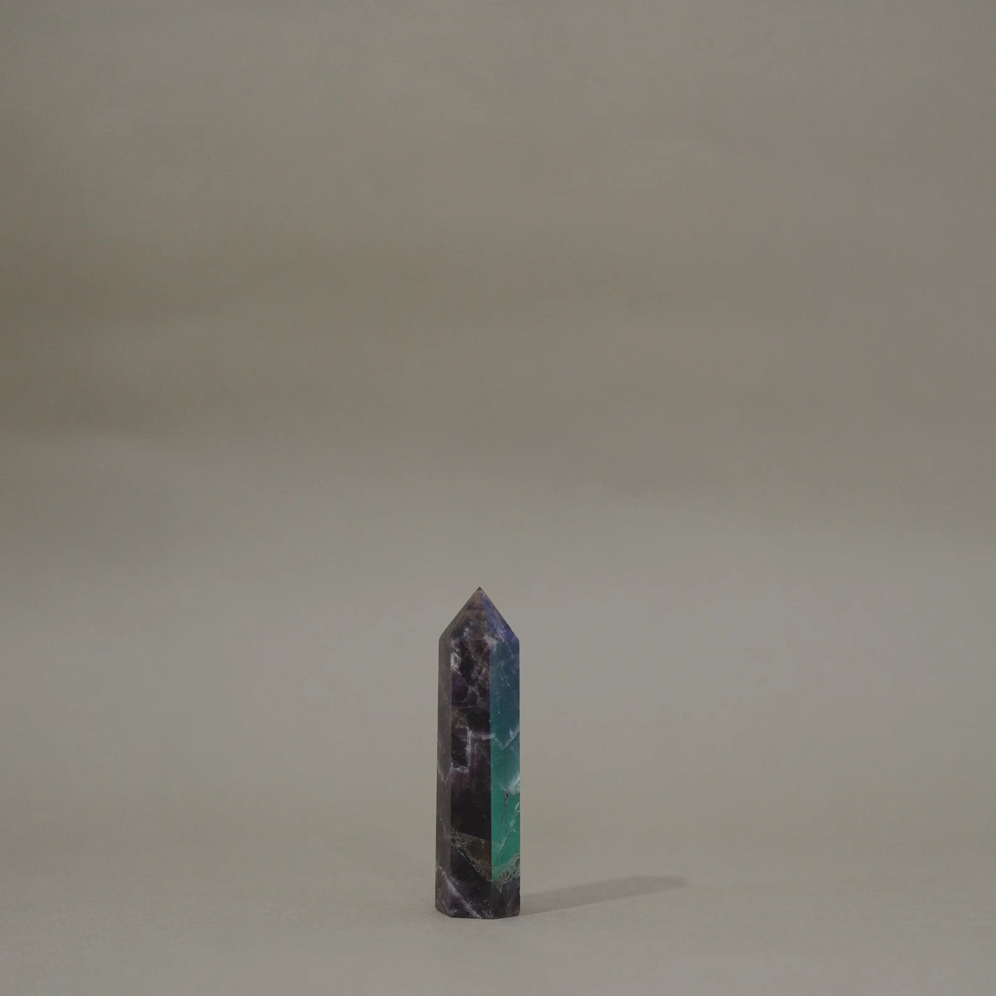 A small polished dream amethyst tower with a marbled pattern of dark purple, black, and teal green, standing upright on a neutral beige surface against a matching beige background.