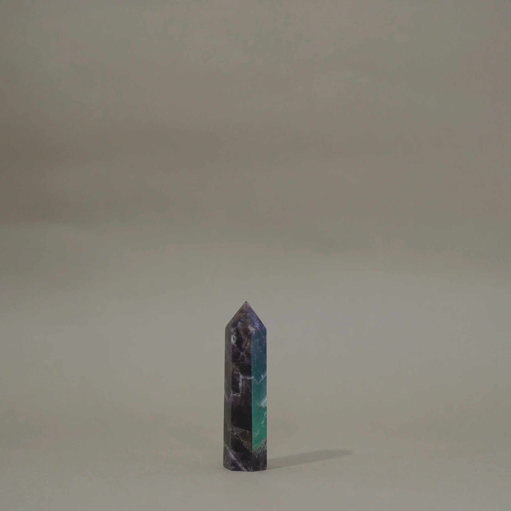 A small polished dream amethyst tower with a marbled pattern of dark purple, black, and teal green, standing upright on a neutral beige surface against a matching beige background.
