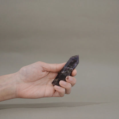 A hand holding a polished dark purple dream amethyst tower against a plain taupe background.