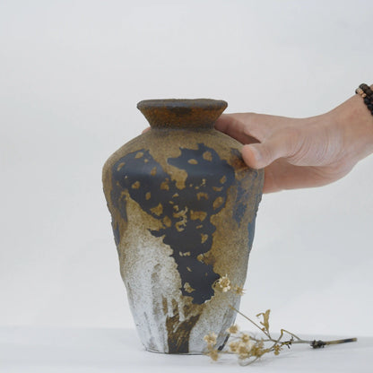 A handmade ceramic vase with a textured, mottled glaze in earthy brown, black, and white, held by a person's hand against a plain white background.