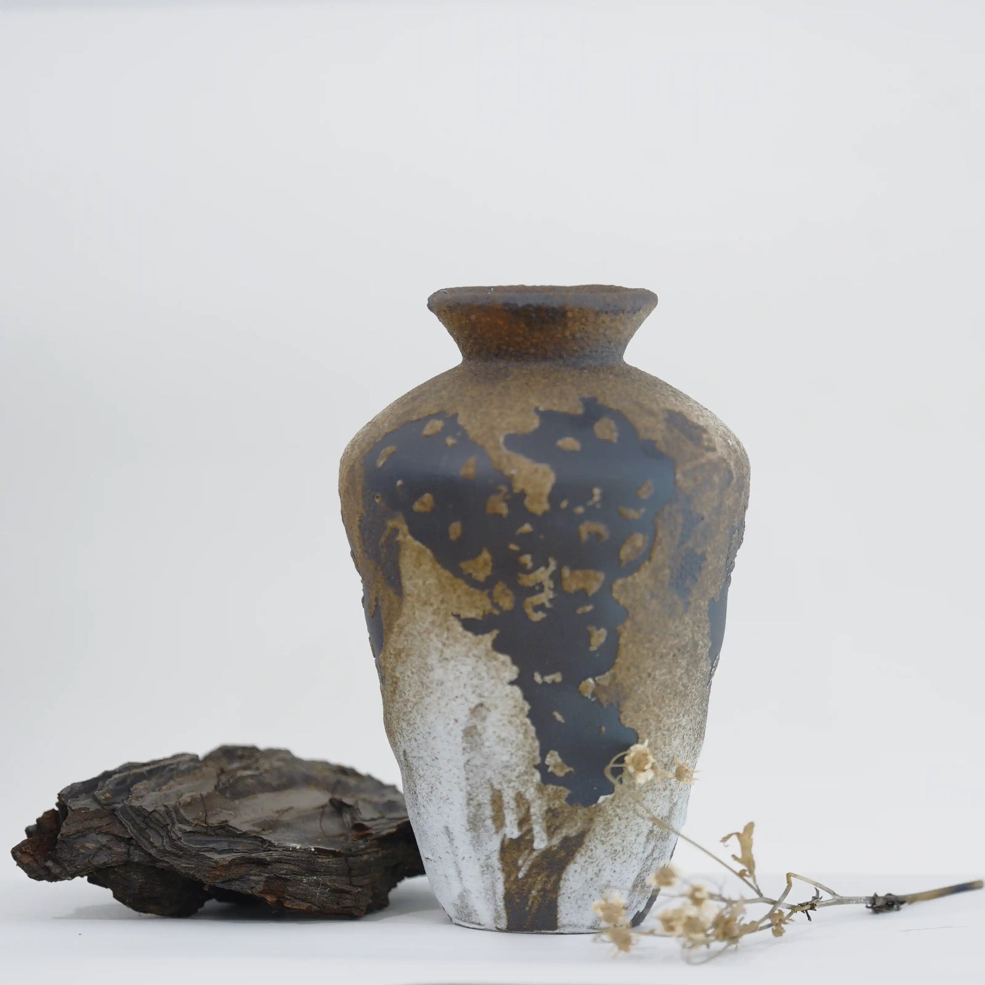 A ceramic vase with a textured, metallic gold and dark brown patina finish, styled on a white surface with a piece of driftwood and dried flowers.