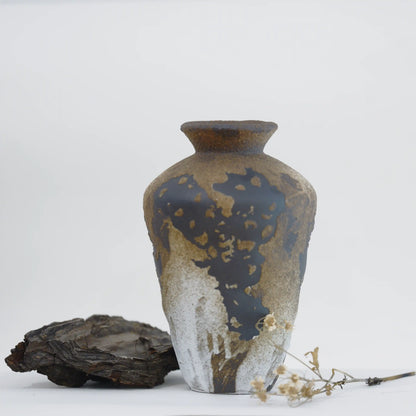 A ceramic vase with a textured, metallic gold and dark brown patina finish, styled on a white surface with a piece of driftwood and dried flowers.