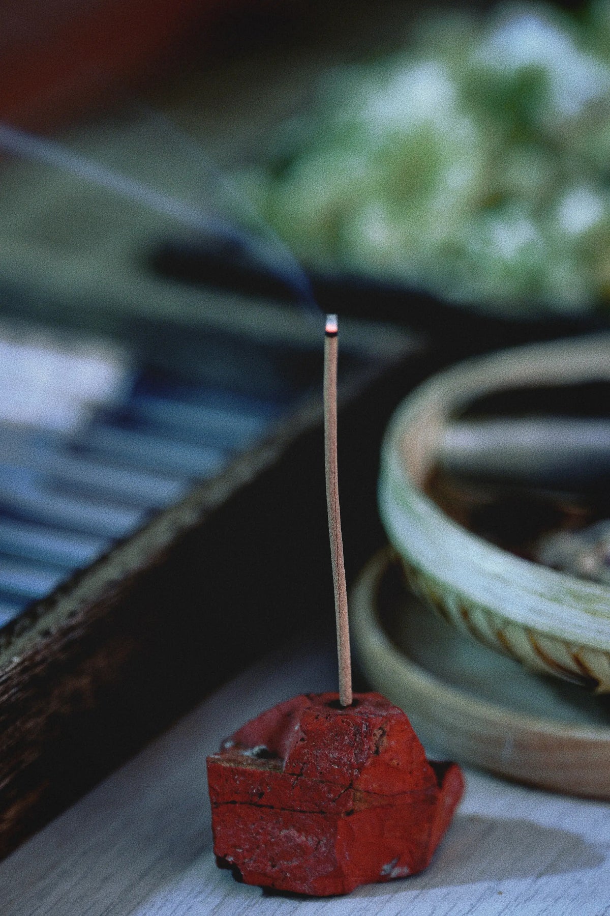 A single incense stick burns in a red ceramic holder on a tabletop, with a shallow dish and blurred objects in the background.