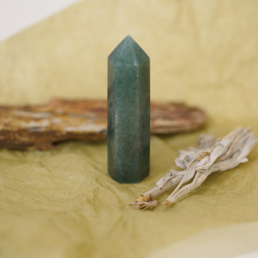 A green aventurine tower stands upright on a crumpled beige surface, with a blurred brown piece of wood and a dried white plant in the background.