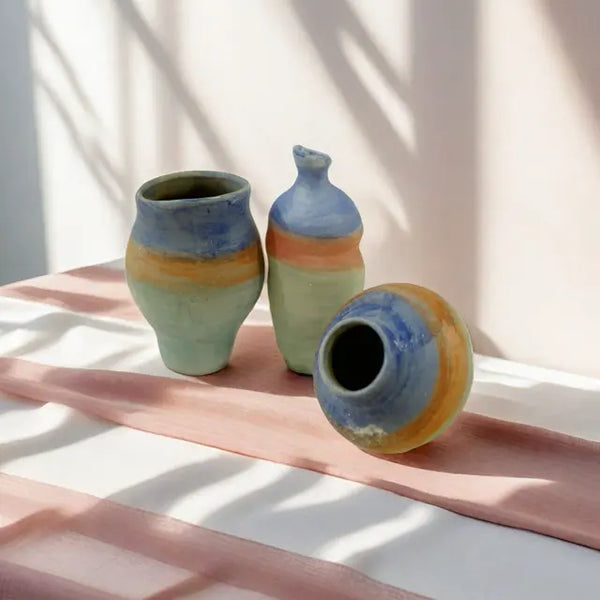 Three artisan ceramic vases with colorful bands, resting on a soft pink cloth, illuminated by gentle shadows.